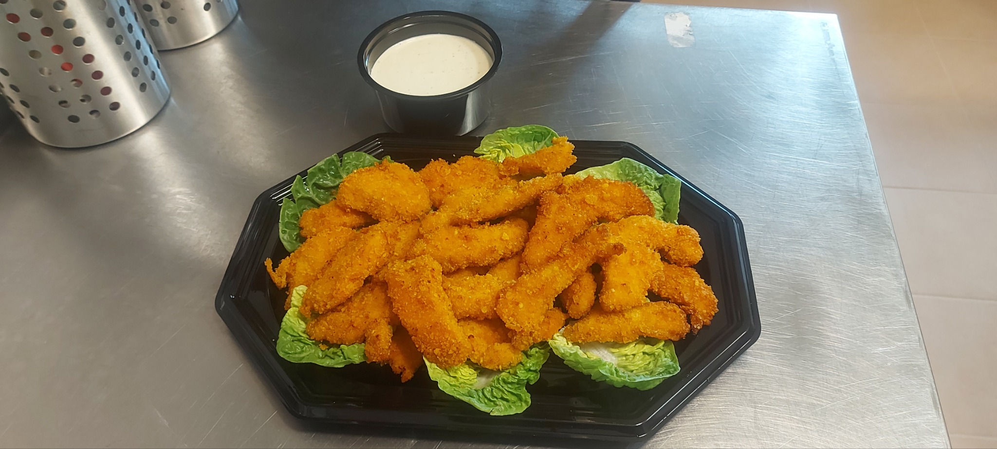 Porcja panierowanych filetów z kurczaka ułożonych na liściach sałaty, podana na czarnym, plastikowym talerzu obok pojemnika z sosem, na metalowym blacie.