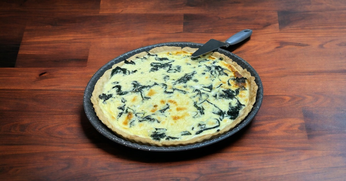Quiche ze szpinakiem w okrągłej formie na drewnianym stole, z łopatką do ciasta, widok z góry.