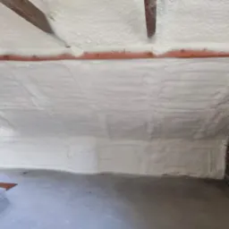 Poddasze z ukończoną izolacją natryskową, widoczne drewniane belki stropowe i fragment ściany z cegły.