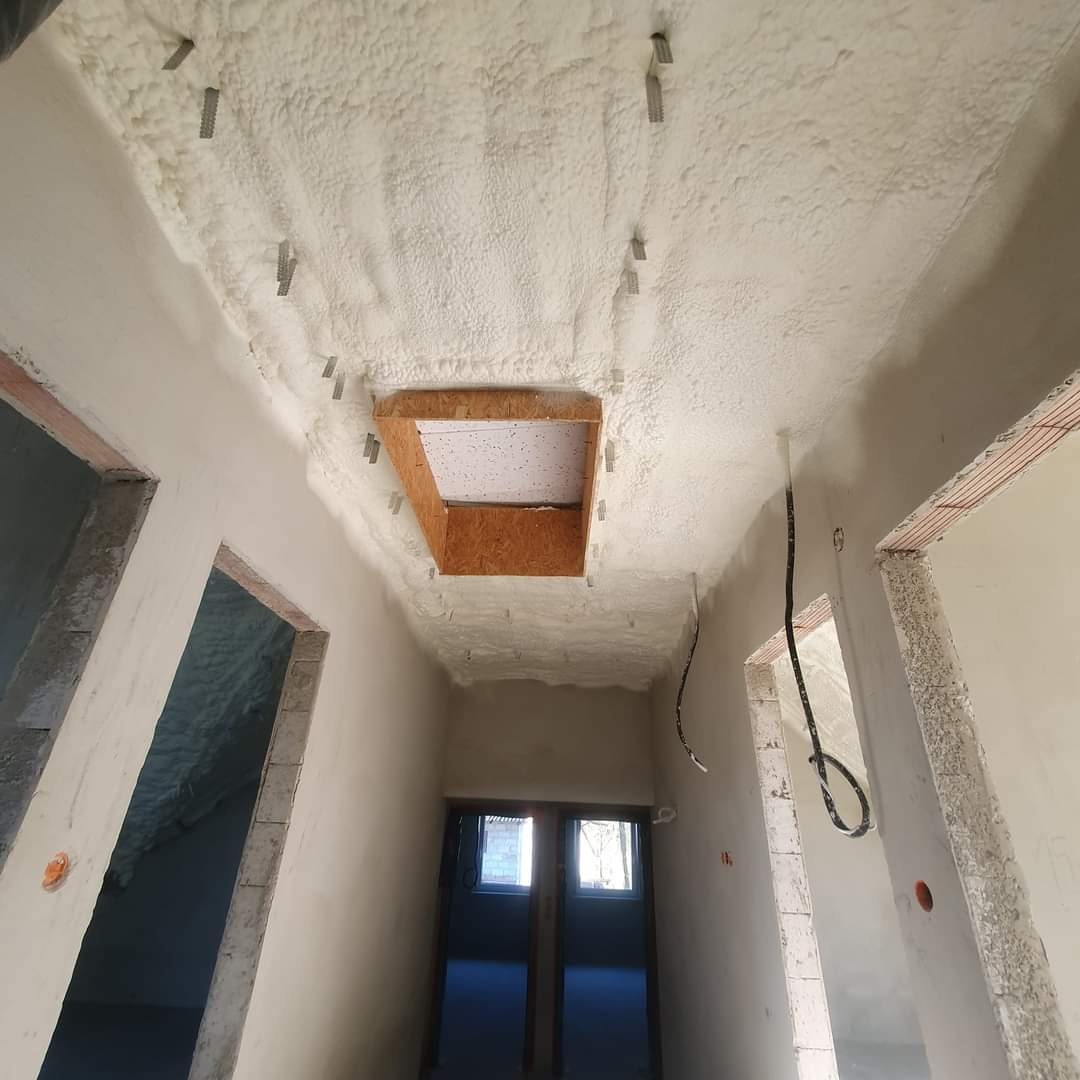 Korytarz w trakcie prac wykończeniowych: sufit pokryty grubą warstwą izolacji, widoczne mocowania, otwór na poddasze z drewnianą obudową, ściany w trakcie tynkowania.