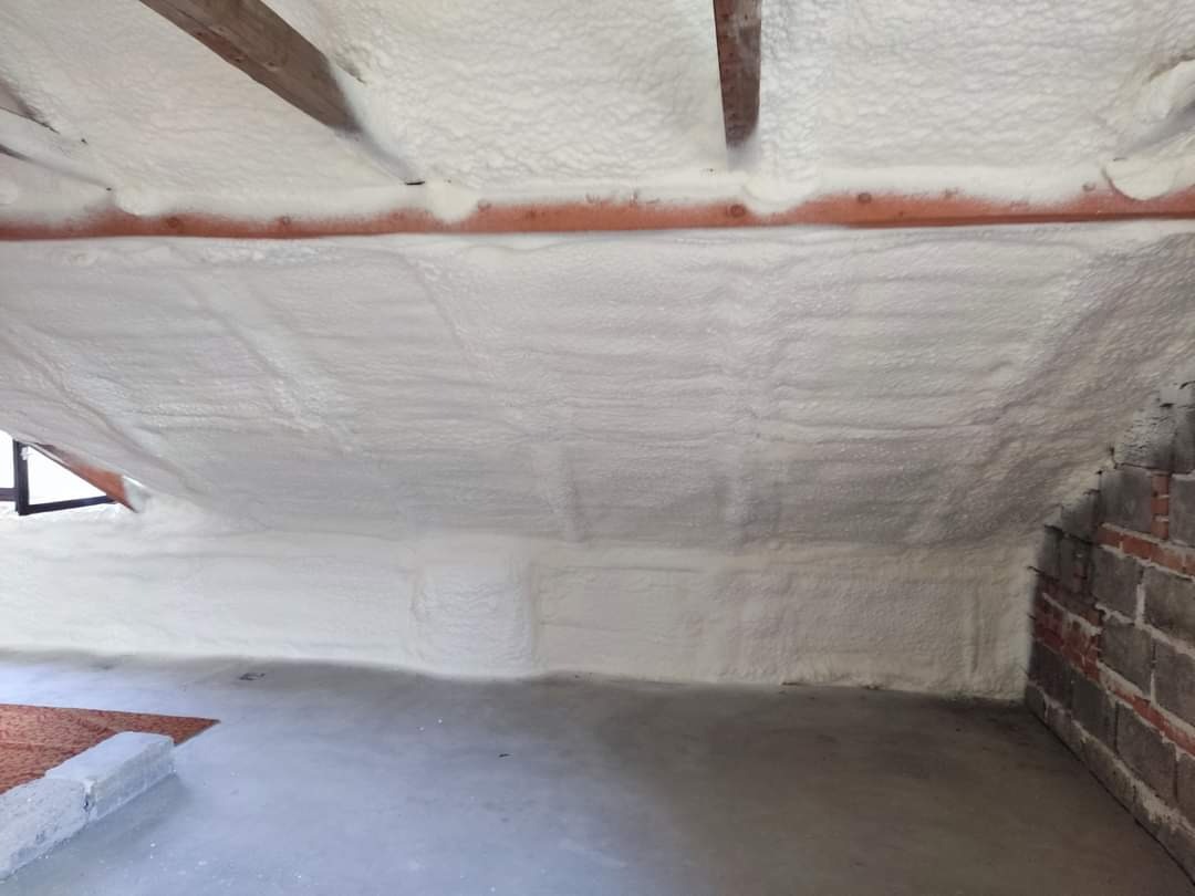 Poddasze z ukończoną izolacją natryskową, widoczne drewniane belki stropowe i fragment ściany z cegły.