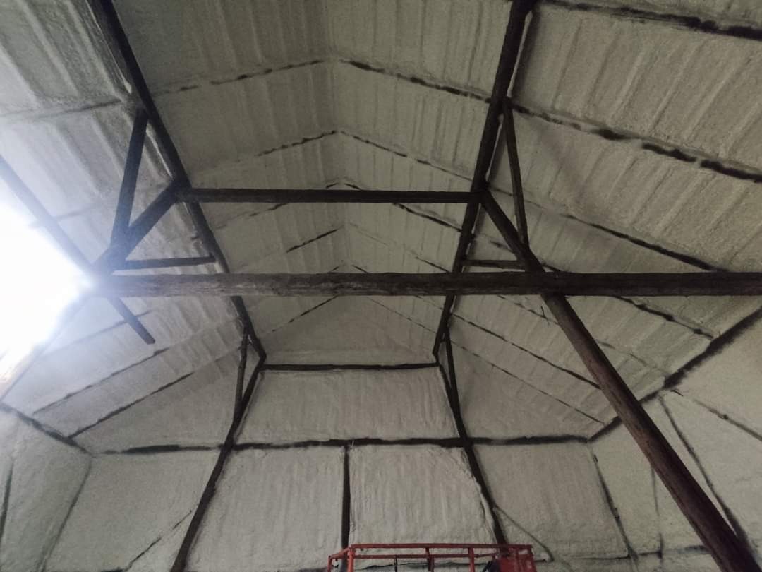 Wnętrze poddasza z drewnianą konstrukcją pokryte warstwą izolacyjną, widoczne nierówności powierzchni i elementy rusztowania w dolnej części kadru.