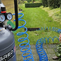 Ogrody Toki Tomasz Słaby - Sprężarka powietrza z niebieskim wężem spiralnym na tle zadbanego ogrodu z trawnikiem i krzewami, sugerująca instalację systemu nawadniania.