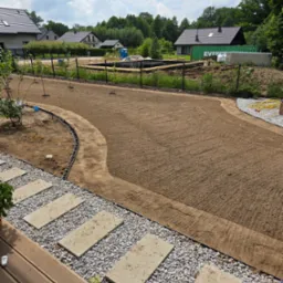 Przygotowany teren pod trawnik, widoczne ścieżki z kamieni i rabaty. W tle domy i drzewa, co sugeruje prace w ogrodzie przydomowym.