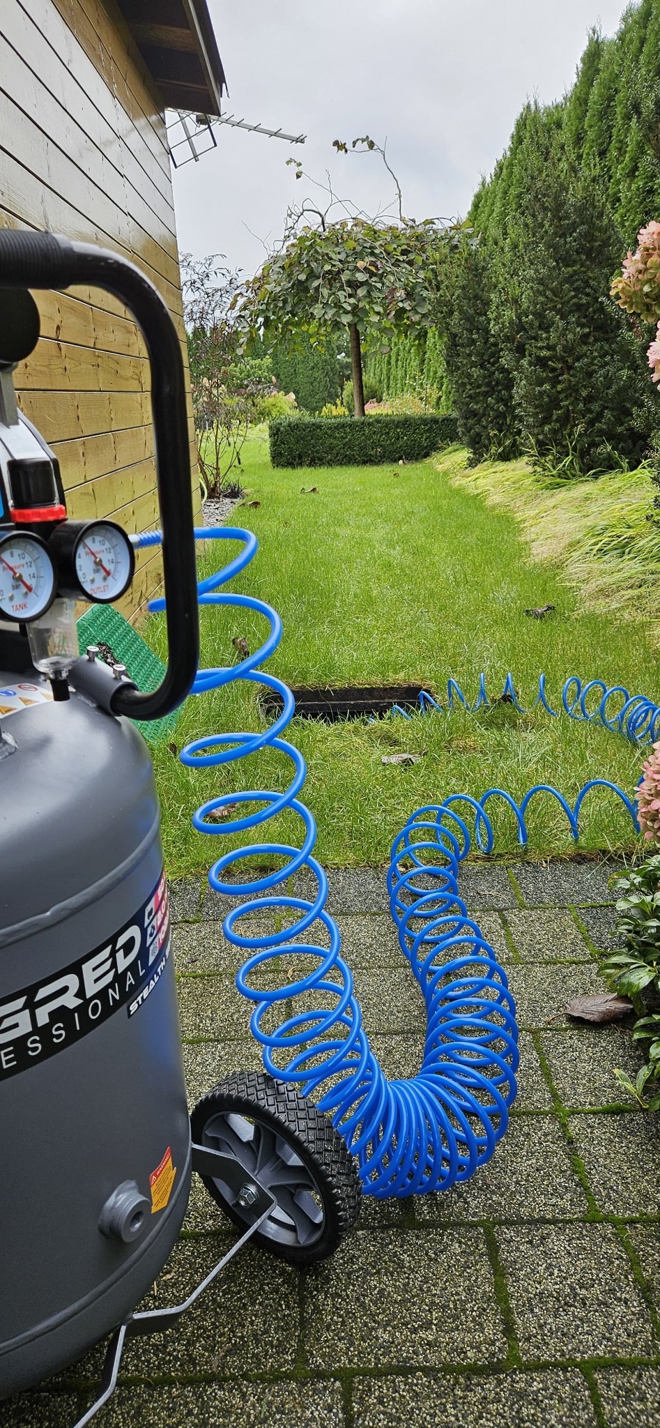 Sprężarka powietrza z niebieskim wężem spiralnym na tle zadbanego ogrodu z trawnikiem i krzewami, sugerująca instalację systemu nawadniania.