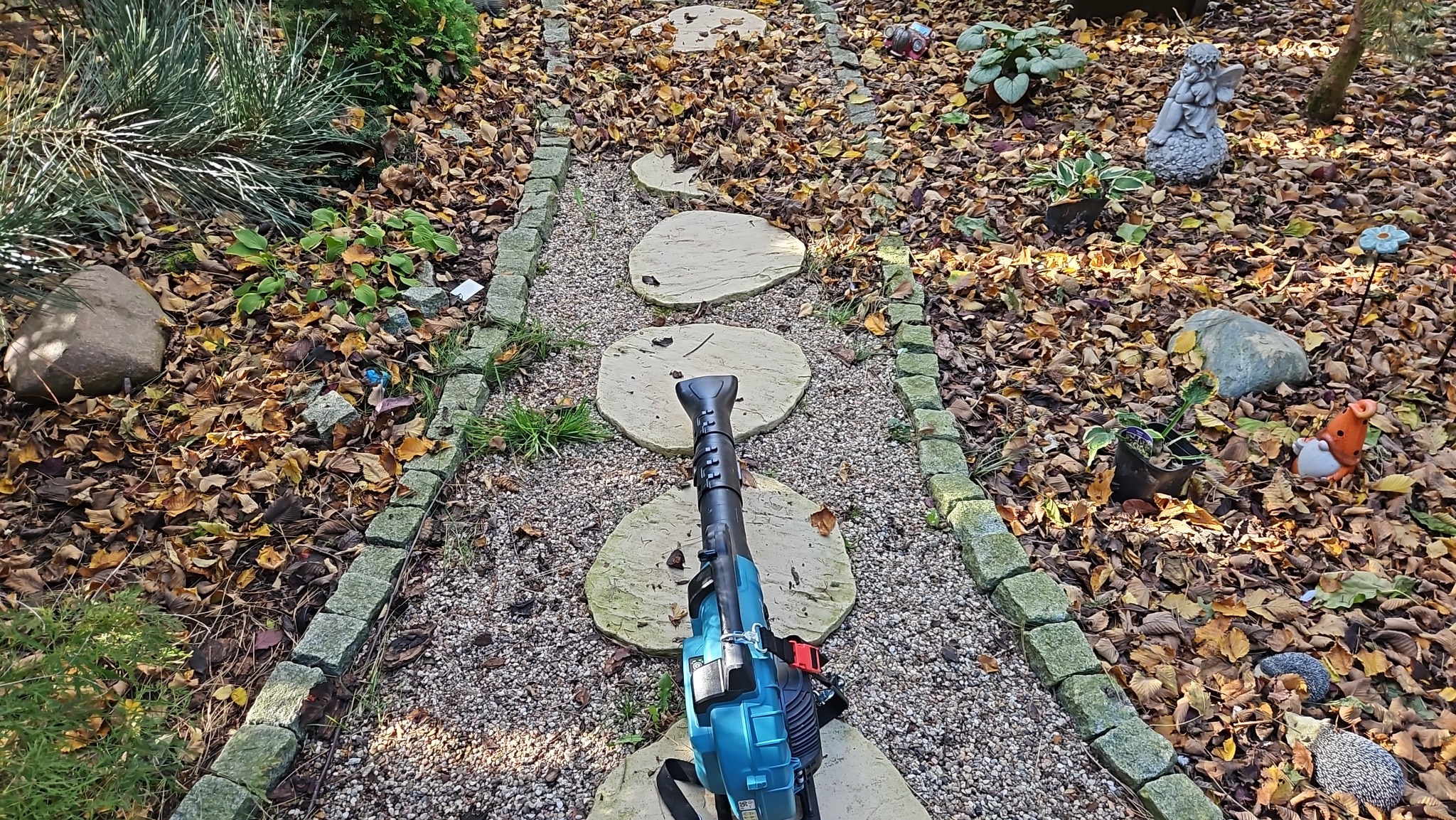 Niebieski odkurzacz do liści leży na ścieżce z kamiennych płyt w ogrodzie pełnym opadłych liści, obok widoczne dekoracje ogrodowe, takie jak figurka anioła i krasnala.