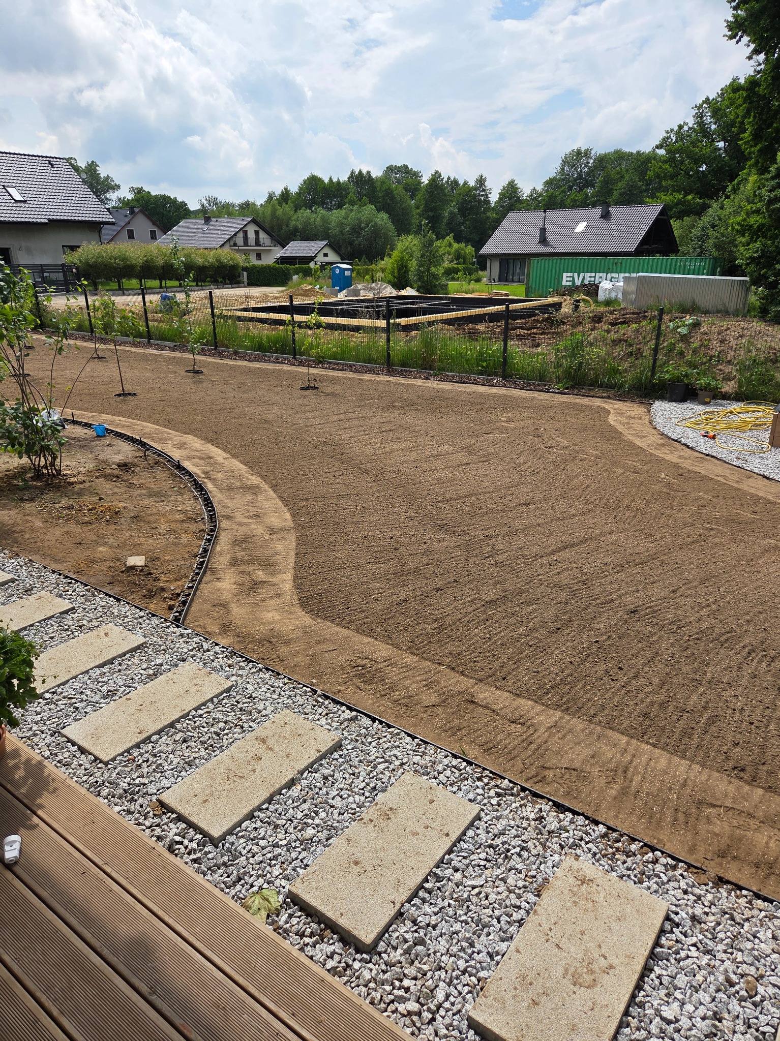 Przygotowany teren pod trawnik, widoczne ścieżki z kamieni i rabaty. W tle domy i drzewa, co sugeruje prace w ogrodzie przydomowym.