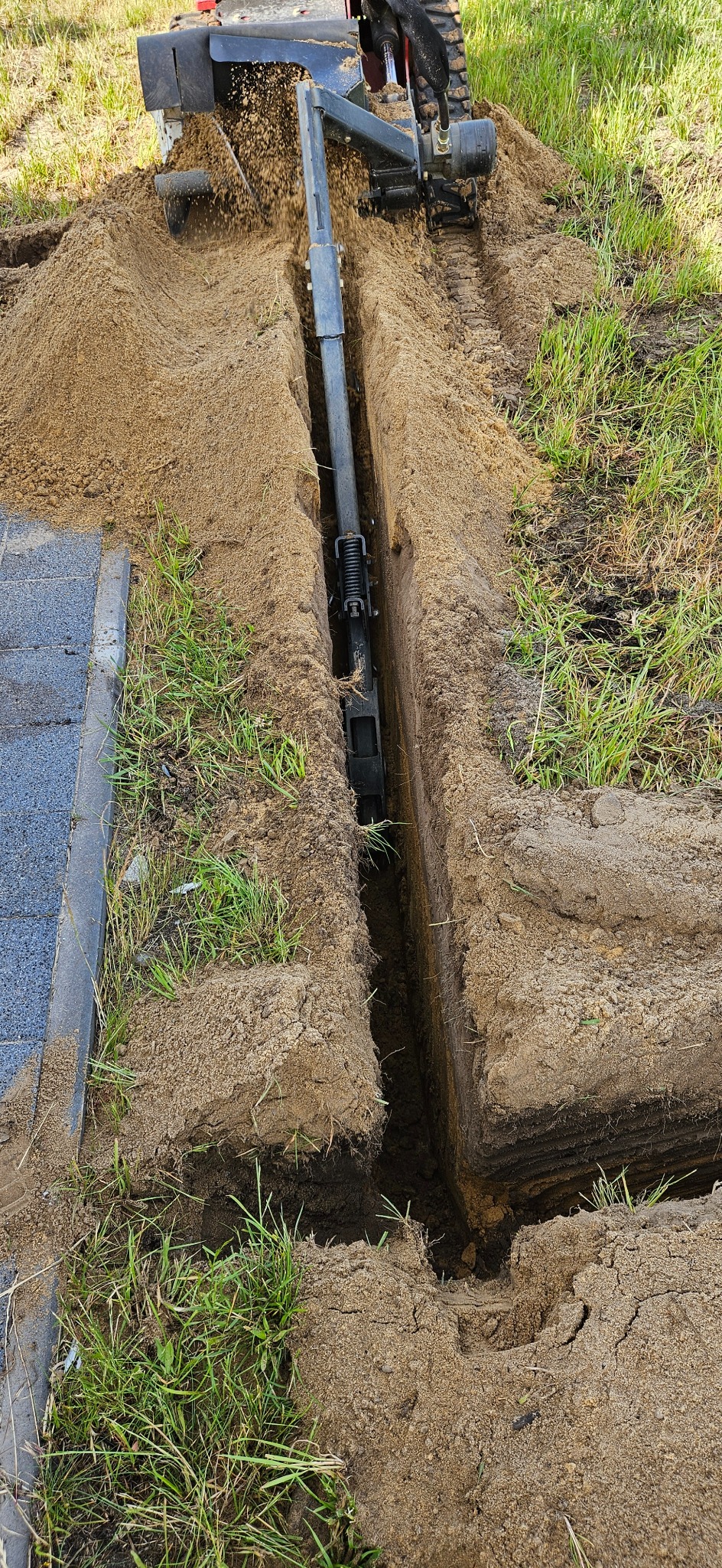 Maszyna wykopuje wąski rów w ziemi obok kostki brukowej, przygotowując teren pod instalację. W tle trawa i świeżo wykopana ziemia.