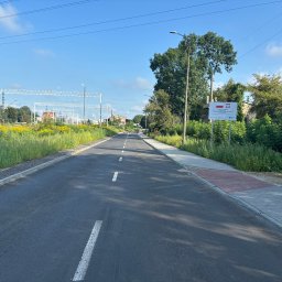 GENERAL DEVELOPMENT SPÓŁKA Z OGRANICZONĄ ODPOWIEDZIALNOŚCIĄ - Nowo wybudowana asfaltowa droga z widocznymi liniami oddzielającymi pasy ruchu, chodnikiem z kostki brukowej po prawej stronie, słupami trakcji kolejowej w tle i tablicą informacyjną po prawej...