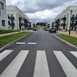 GENERAL DEVELOPMENT SPÓŁKA Z OGRANICZONĄ ODPOWIEDZIALNOŚCIĄ - Nowa, asfaltowa droga osiedlowa z białymi pasami przejścia dla pieszych, otoczona nowoczesnymi budynkami mieszkalnymi z balkonami i zadbanymi trawnikami.