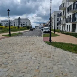 Ulica wyłożona szarą kostką brukową w nowym osiedlu mieszkaniowym, widoczne lampy uliczne i chodnik z kostki czerwonej.