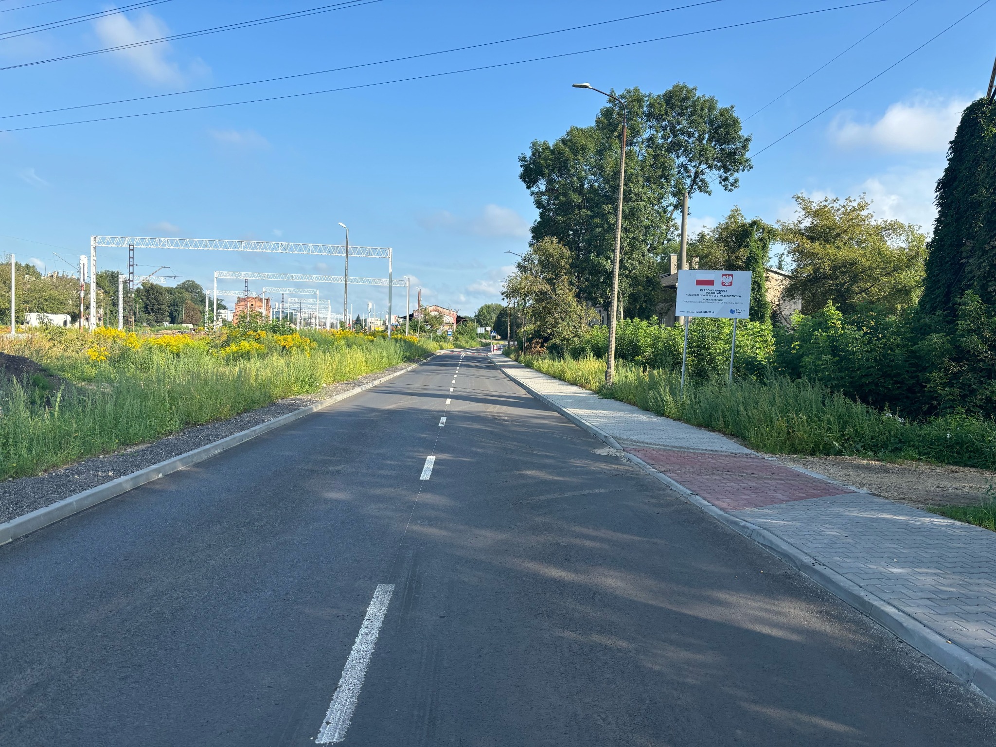 Nowo wybudowana asfaltowa droga z widocznymi liniami oddzielającymi pasy ruchu, chodnikiem z kostki brukowej po prawej stronie, słupami trakcji kolejowej w tle i tablicą informacyjną po prawej...
