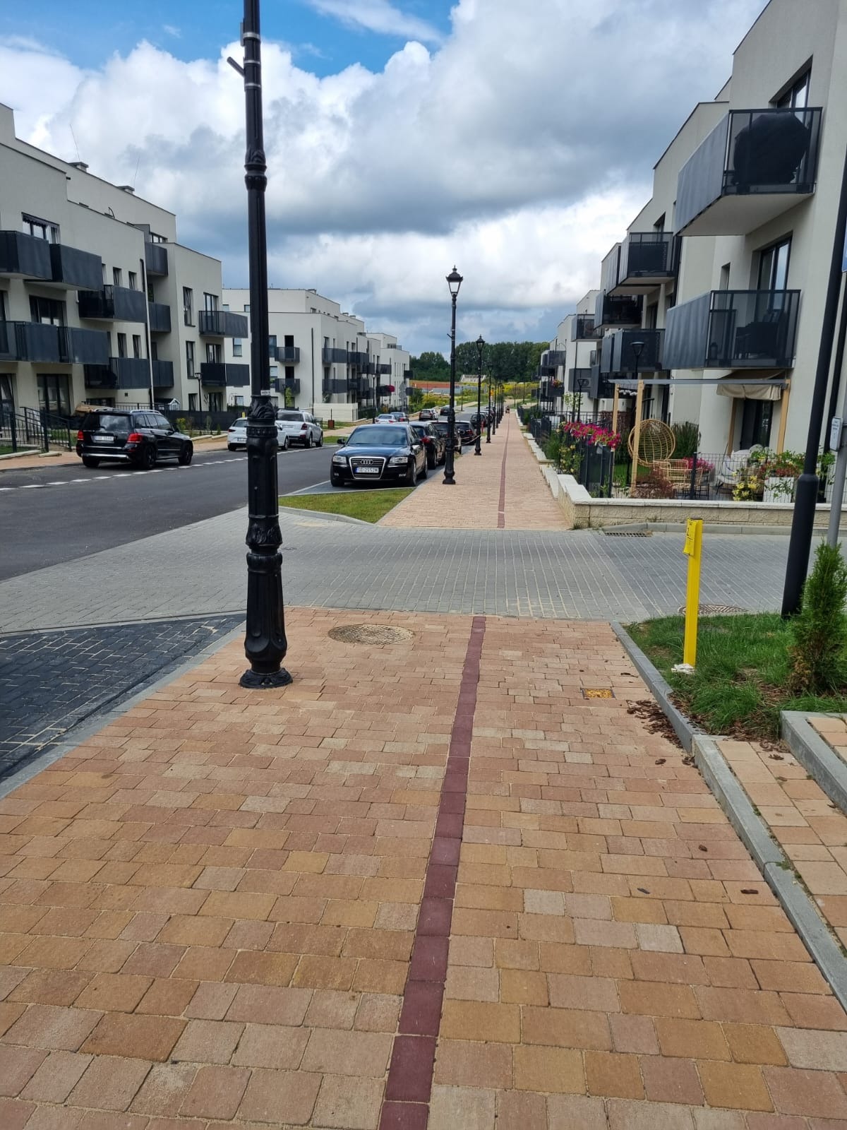 Nowoczesne osiedle z chodnikiem z kostki brukowej w odcieniach beżu i brązu, oddzielonego ciemniejszym pasem, oświetlonym stylowymi latarniami.