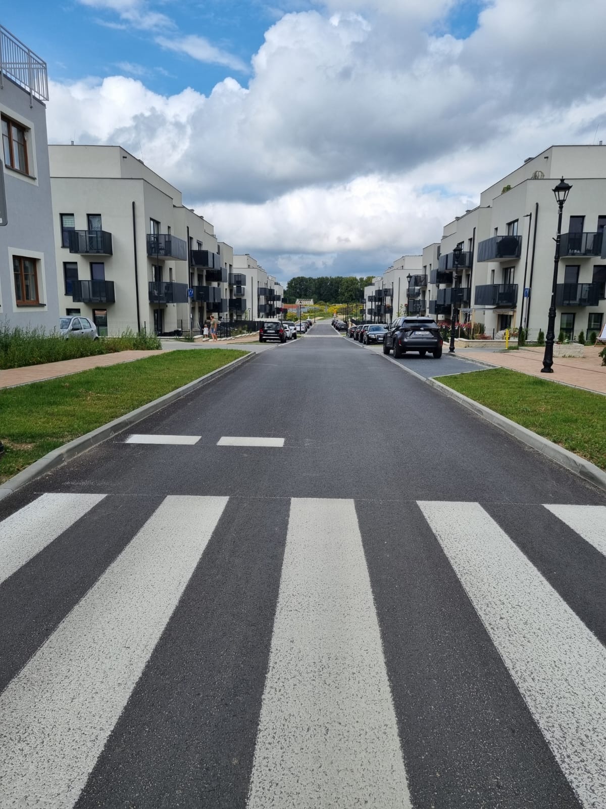 Nowa, asfaltowa droga osiedlowa z białymi pasami przejścia dla pieszych, otoczona nowoczesnymi budynkami mieszkalnymi z balkonami i zadbanymi trawnikami.