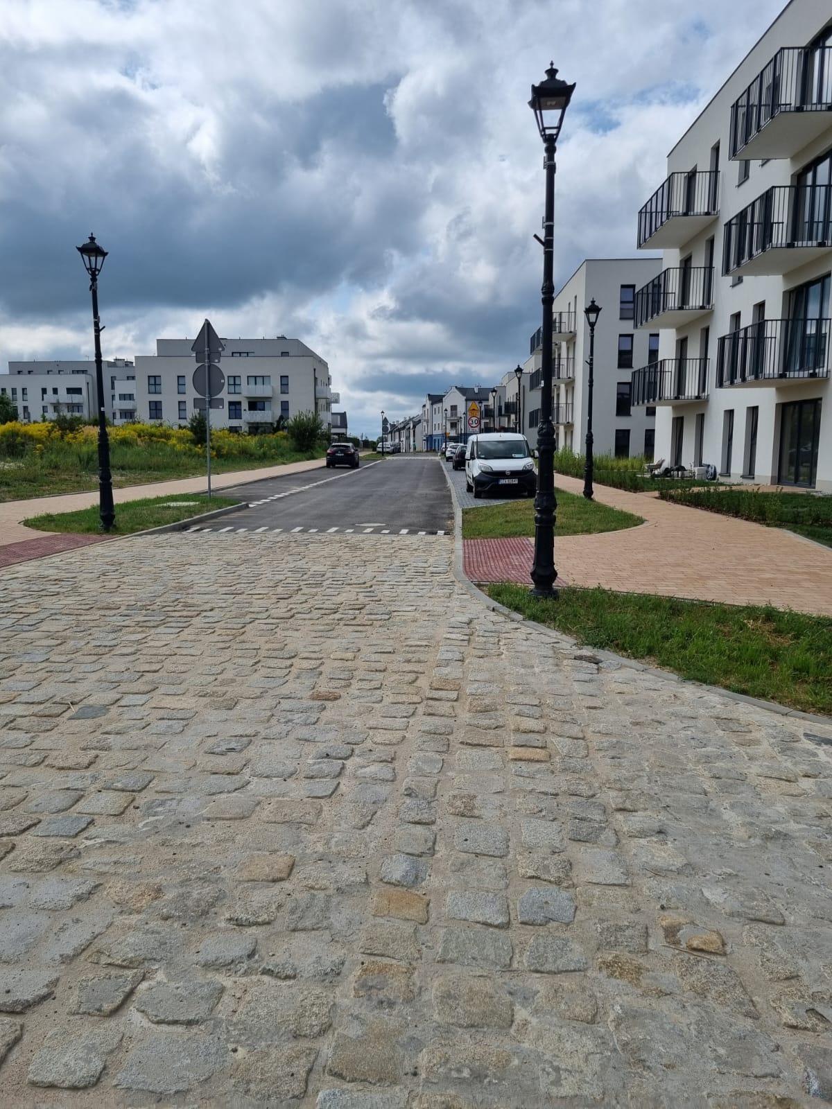 Ulica wyłożona szarą kostką brukową w nowym osiedlu mieszkaniowym, widoczne lampy uliczne i chodnik z kostki czerwonej.