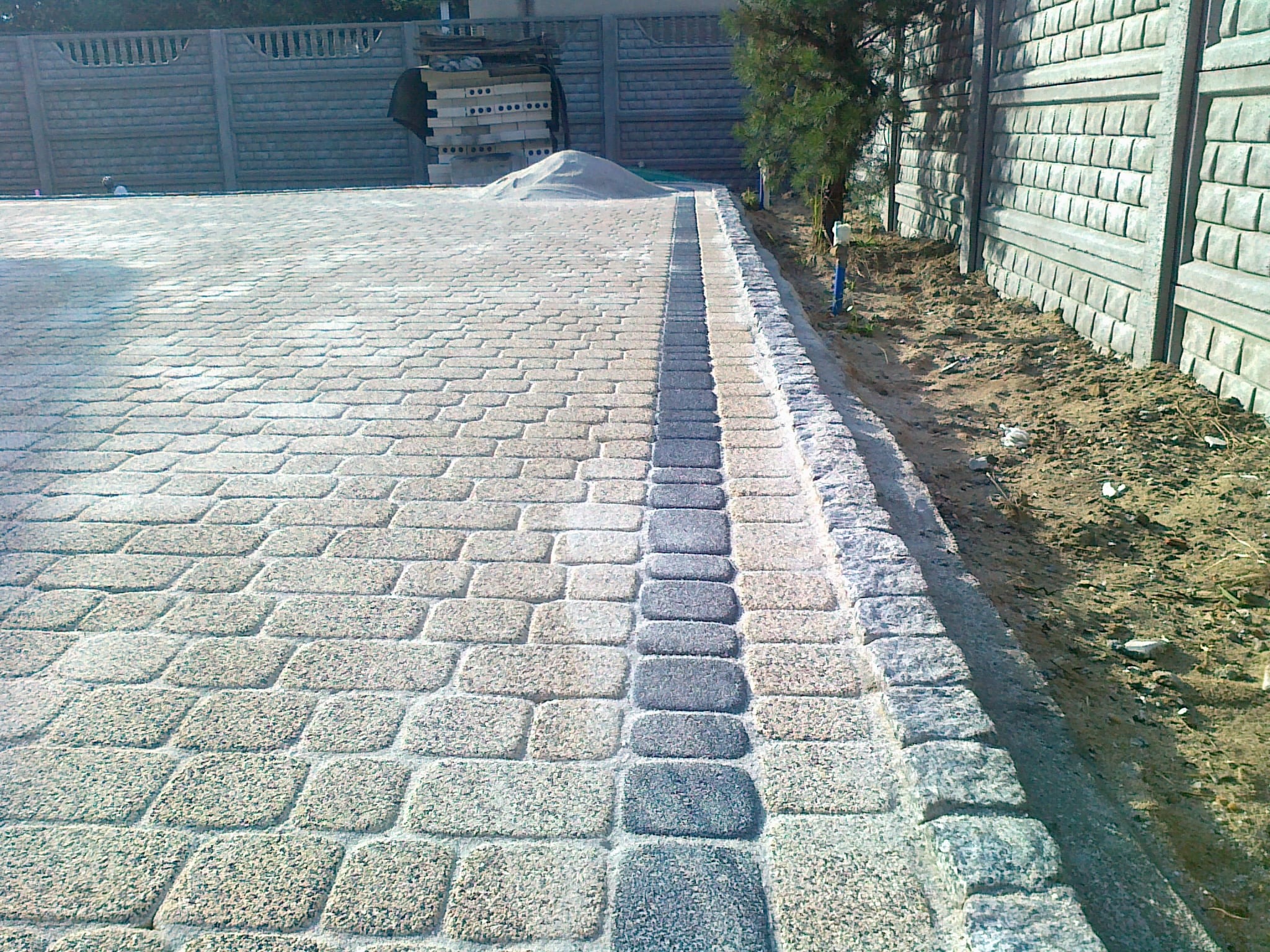 Ułożona kostka brukowa w dwóch odcieniach szarości, z ciemniejszym pasem pośrodku, otoczona betonowym ogrodzeniem i zielenią.