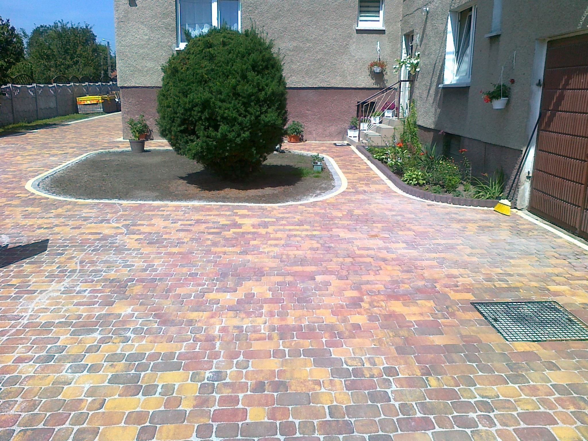 Kolorowa kostka brukowa ułożona przed domem z zielonym trawnikiem i ozdobnymi roślinami w donicach, widoczna studzienka kanalizacyjna z metalową kratką.