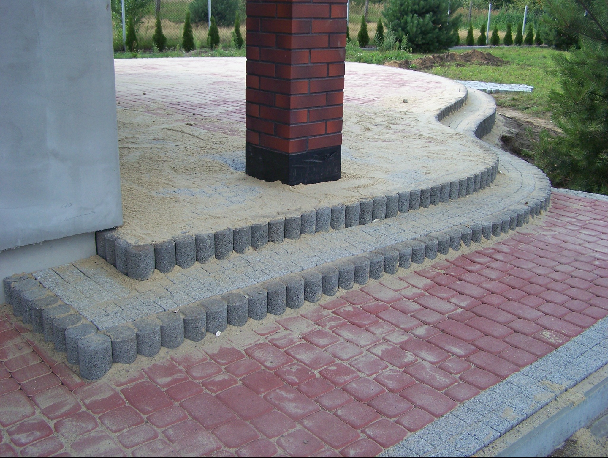Czerwona kostka brukowa ułożona wzorem cegły, z obrzeżami z szarej kostki w kształcie walca, prowadząca do schodów z szarej kostki i ceglanej kolumny, całość zasypana piaskiem.