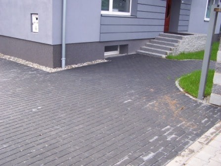 Ułożona, ciemnoszara kostka brukowa przed nowoczesnym domem z szarą elewacją, widoczne schody wejściowe i fragment trawnika.