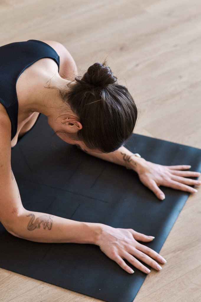 Kobieta z tatuażami na przedramionach wykonuje pozycję sfinksa na czarnej macie do jogi, włosy upięte w kok, widoczny parkiet.