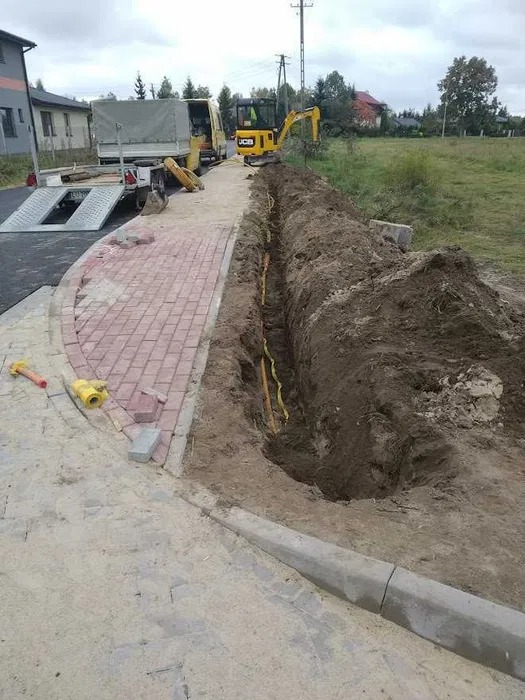Żółte rury w wykopie wzdłuż chodnika z kostki, mini koparka JCB i ciężarówka z lawetą w tle, widoczne narzędzia i świeżo ułożona kostka chodnikowa.