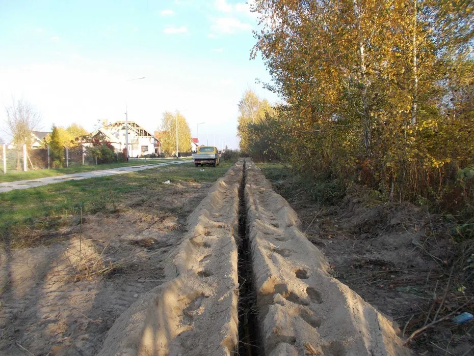 Długi, prosty wykop w ziemi, prawdopodobnie pod instalacje, biegnący wzdłuż drogi wiejskiej z widocznym samochodem w oddali i zabudowaniami w tle.
