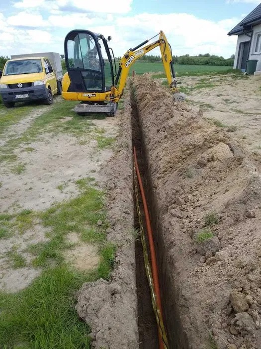 Żółta mini koparka JCB obok wykopanego rowu z rurą w ziemi, na tle trawnika i żółtego samochodu dostawczego.