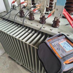 Ampirion Kacper Korzeniowski - Transformator z podłączonym miernikiem Sonel MIC-2501. Widoczne przewody z krokodylkami, brązowe izolatory i metalowa obudowa. Pomiar rezystancji izolacji.