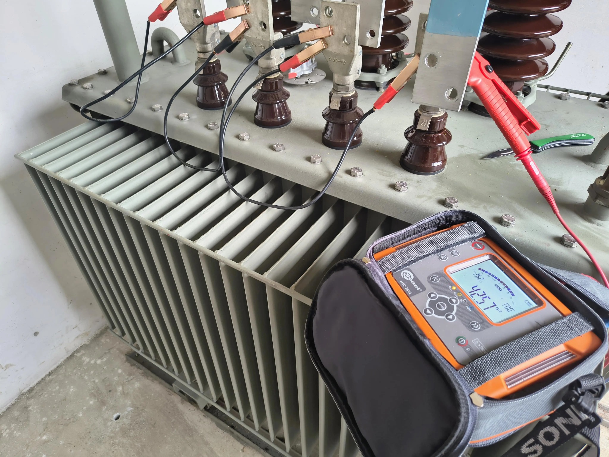 Transformator z podłączonym miernikiem Sonel MIC-2501. Widoczne przewody z krokodylkami, brązowe izolatory i metalowa obudowa. Pomiar rezystancji izolacji.