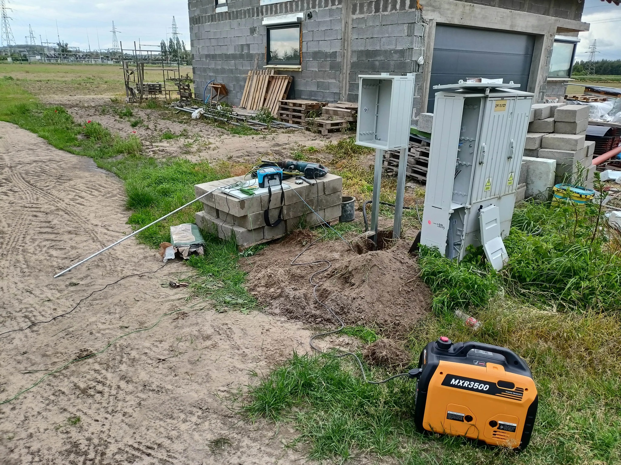 Montaż skrzynki elektrycznej przy nowym budynku. Widoczny agregat prądotwórczy, narzędzia i materiały. W tle słupy wysokiego napięcia.