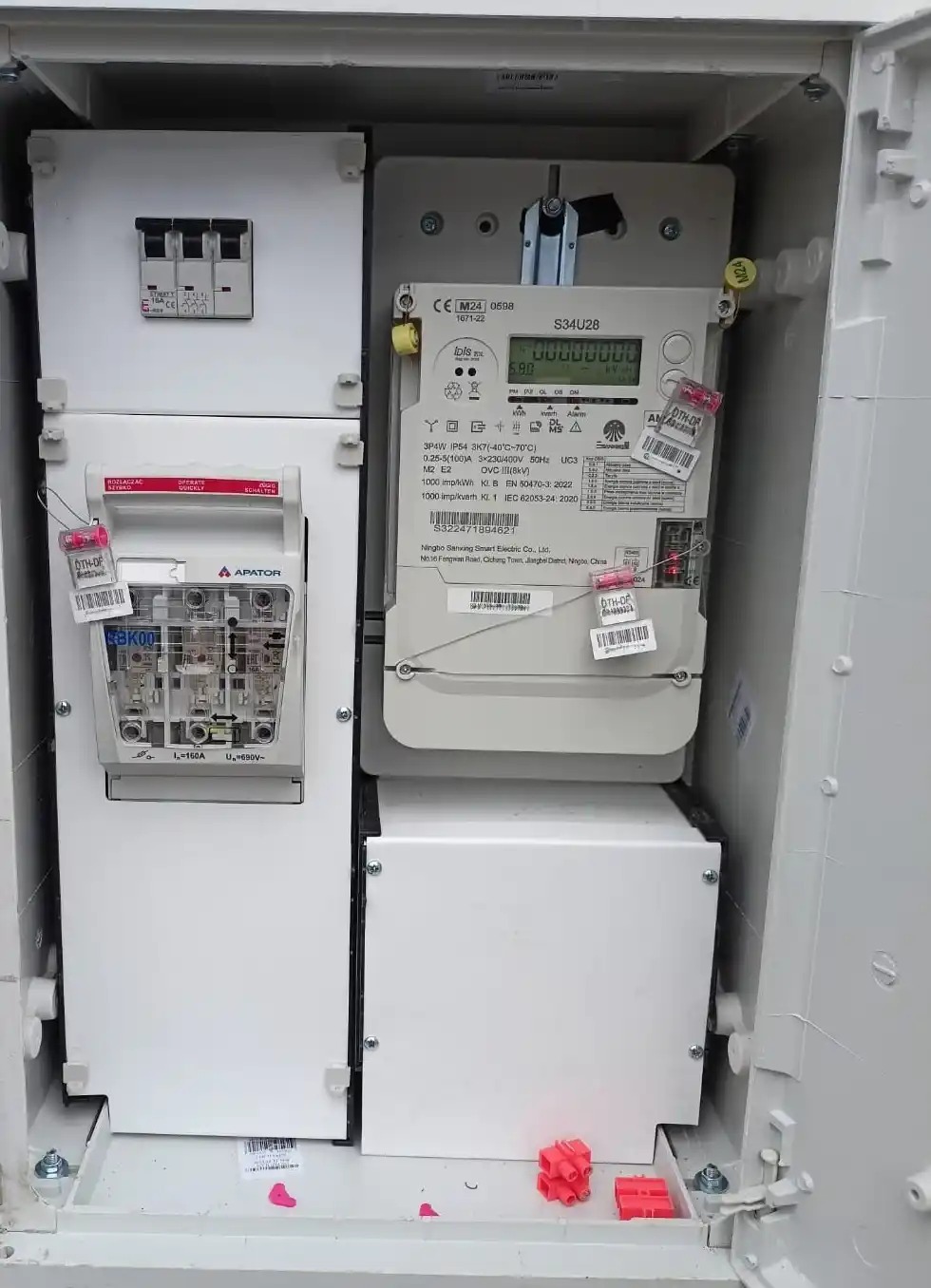 Zamknięta skrzynka z zamontowanymi licznikami energii elektrycznej i zabezpieczeniami. Widoczne plombowania i etykiety identyfikacyjne.