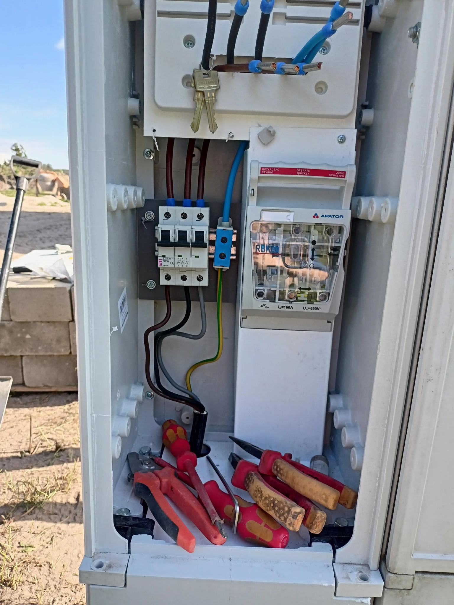 Otwarta skrzynka elektryczna z zainstalowanymi bezpiecznikami, licznikiem i przewodami. Wewnątrz leżą narzędzia: śrubokręty i kombinerki. Widoczne klucze.