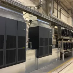 Dwa duże, czarne urządzenia z metalowymi obudowami i kratkami wentylacyjnymi, połączone systemem rur i kanałów wentylacyjnych w przemysłowym pomieszczeniu z betonową podłogą i ścianami.