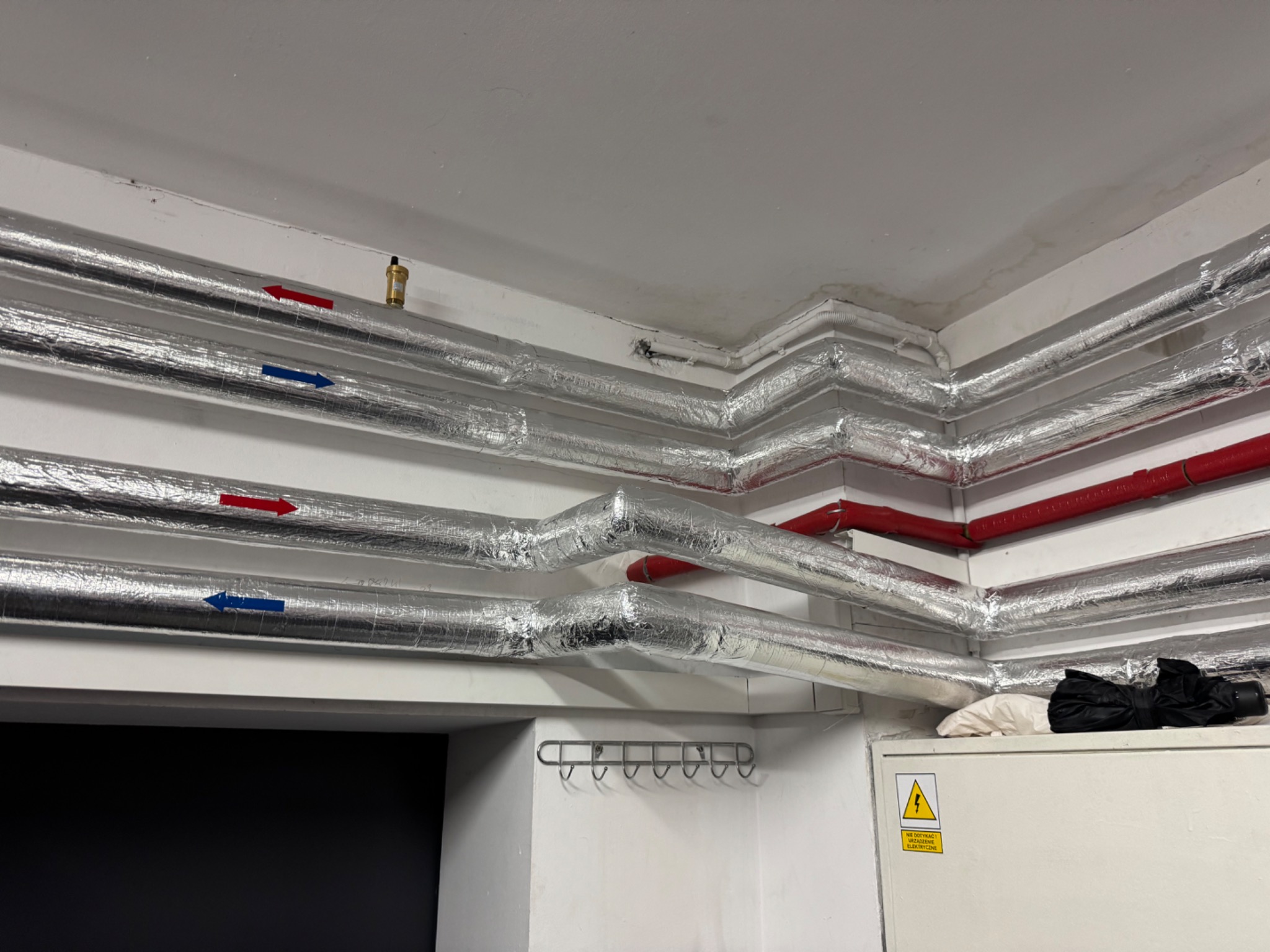 Izolowane rury instalacji grzewczej pod sufitem, z widocznymi strzałkami wskazującymi kierunek przepływu, zamontowane w pomieszczeniu technicznym z wiszącym wieszakiem na ścianie i szafką elektryczną.