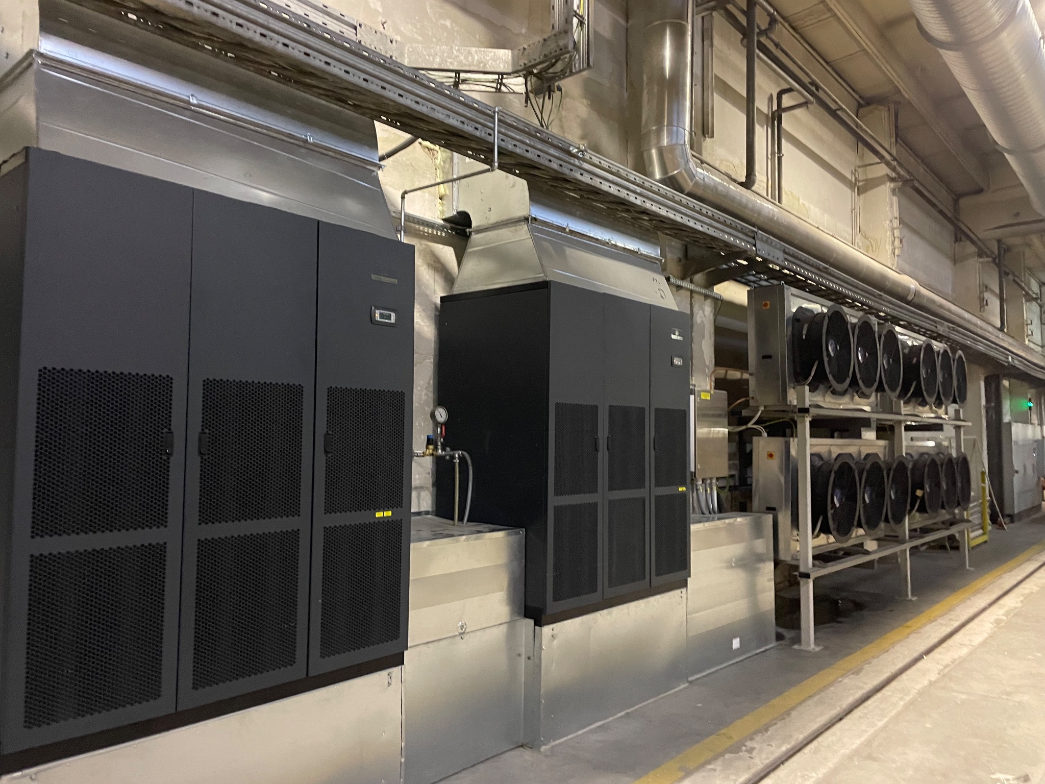 Dwa duże, czarne urządzenia z metalowymi obudowami i kratkami wentylacyjnymi, połączone systemem rur i kanałów wentylacyjnych w przemysłowym pomieszczeniu z betonową podłogą i ścianami.