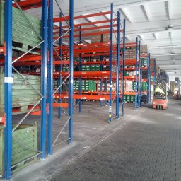 mLog24 - Wnętrze magazynu z regałami wypełnionymi towarami, widoczny wózek widłowy w oddali.