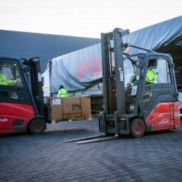 mLog24 - Dwa wózki widłowe Linde transportują kartonowe pudła na paletach do naczepy ciężarówki, pracownicy w odblaskowych kamizelkach, szary budynek magazynowy w tle.