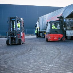 mLog24 - Dwa wózki widłowe Linde na placu manewrowym, jeden z ładunkiem przy naczepie ciężarówki, pracownicy w odblaskowych kamizelkach, szara kostka brukowa.