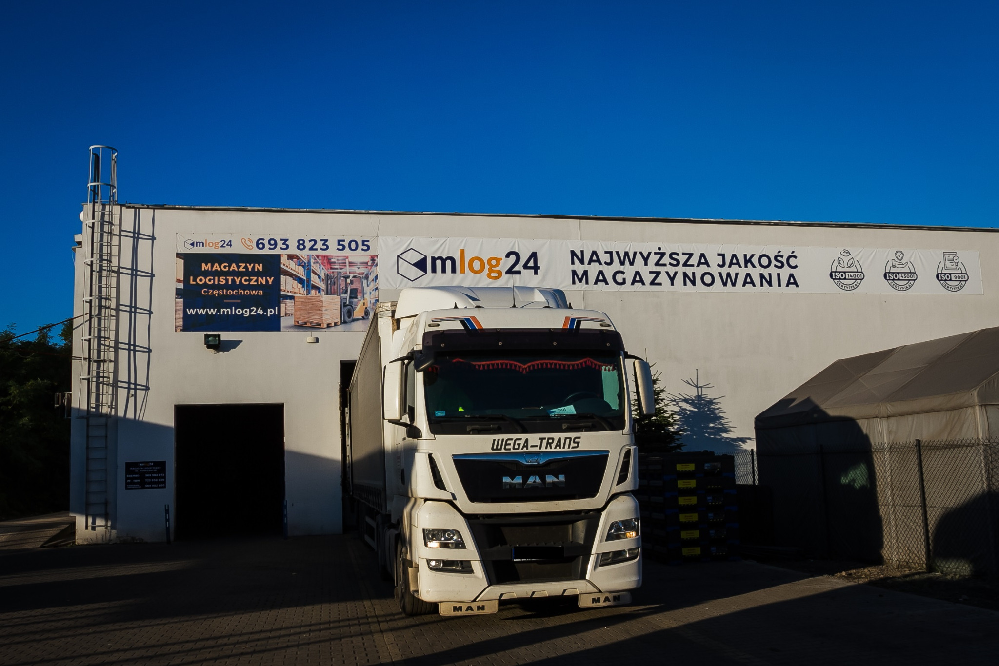 Ciężarówka Wega-Trans MAN wjeżdża do magazynu logistycznego mlog24 w Częstochowie, na budynku reklama i certyfikaty ISO.