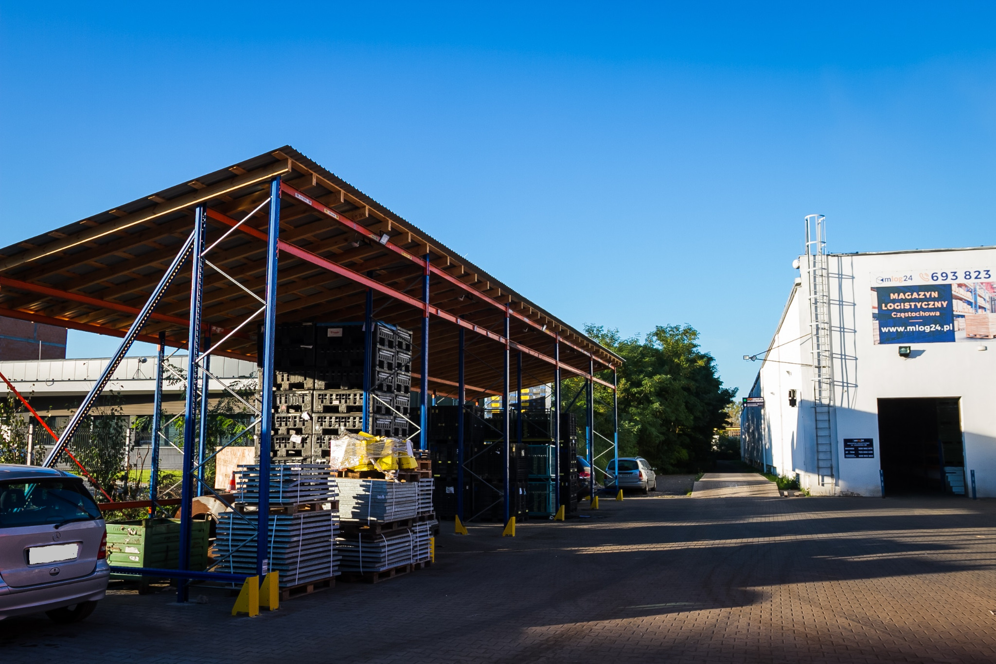 Zadaszona wiata magazynowa z metalową konstrukcją, wypełniona paletami z czarnymi skrzynkami i szarymi płytami, na tle budynku z reklamą magazynu logistycznego w Częstochowie.