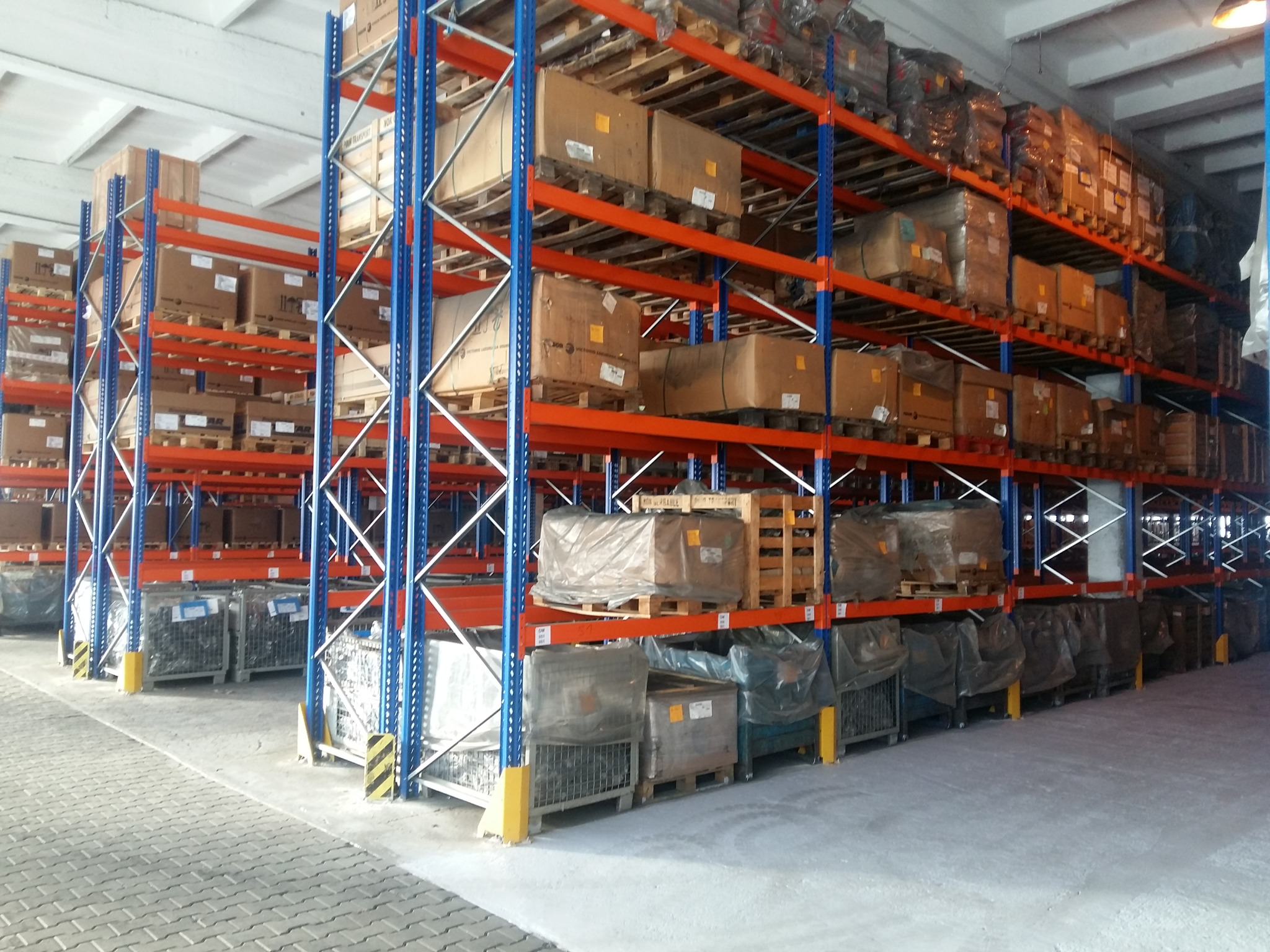 Pełne regały magazynowe z kartonami i metalowymi skrzyniami, widok z perspektywy posadzki magazynu.