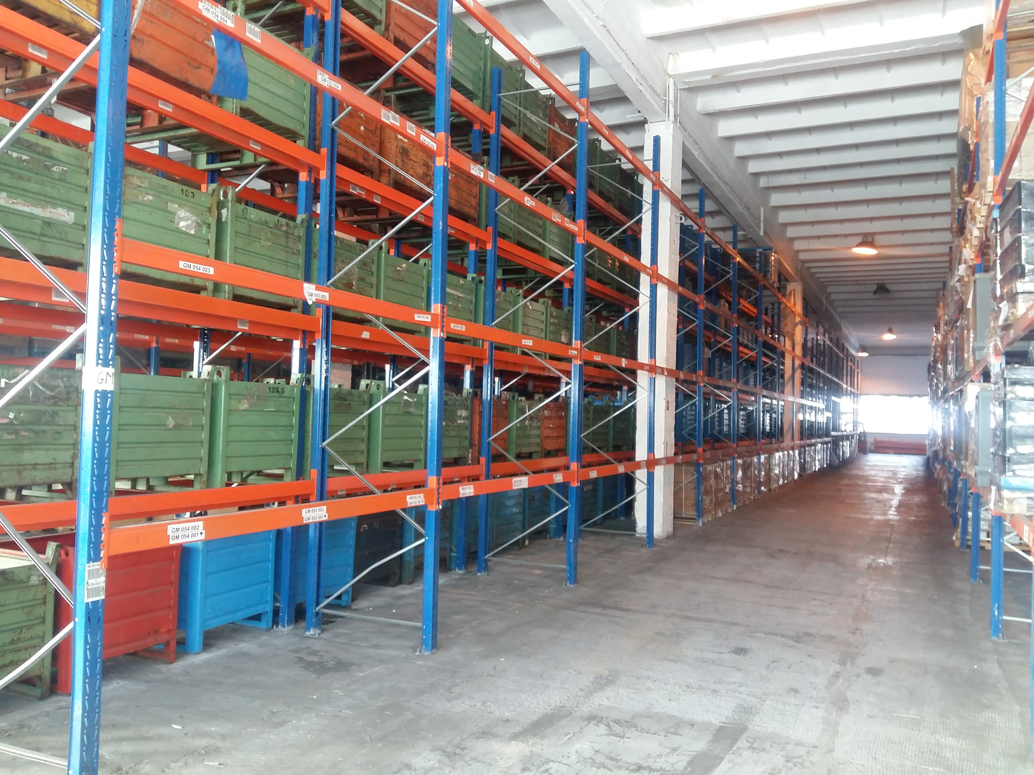 Widok perspektywiczny wnętrza magazynu z wysokimi regałami metalowymi w kolorach pomarańczowym i niebieskim, wypełnionymi metalowymi skrzyniami w odcieniach zieleni, brązu, czerwieni i błękitu,...