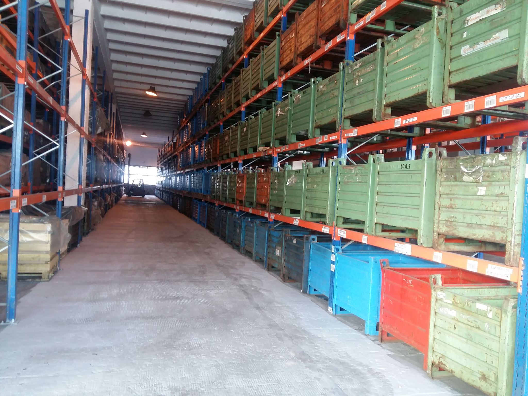 Wnętrze magazynu z regałami wypełnionymi metalowymi skrzyniami w różnych kolorach: zielonym, niebieskim i czerwonym. Widoczny wózek widłowy w oddali.