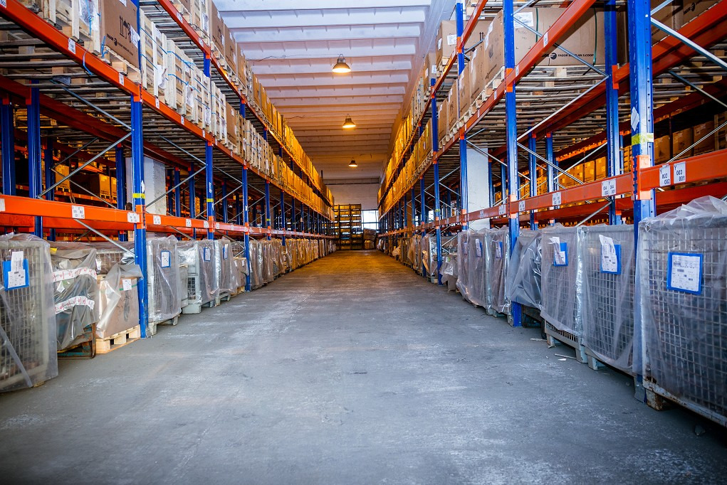 Długi korytarz magazynowy z regałami wypełnionymi kartonami i metalowymi kontenerami zabezpieczonymi folią, oświetlony lampami sufitowymi.