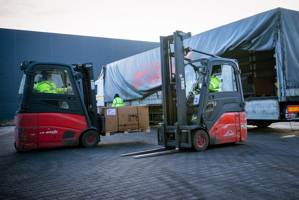 Dwa wózki widłowe Linde transportują kartonowe pudła na paletach do naczepy ciężarówki, pracownicy w odblaskowych kamizelkach, szary budynek magazynowy w tle.