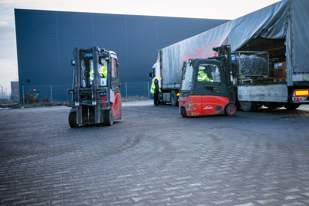 Dwa wózki widłowe Linde na placu manewrowym, jeden z ładunkiem przy naczepie ciężarówki, pracownicy w odblaskowych kamizelkach, szara kostka brukowa.