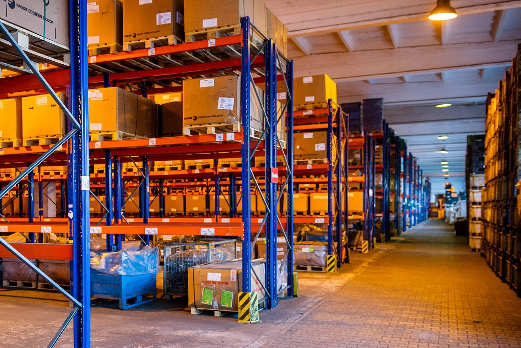 Wnętrze magazynu z wysokimi regałami wypełnionymi kartonami na paletach, oświetlone ciepłym światłem lamp sufitowych, z widoczną perspektywą długiego korytarza między regałami.