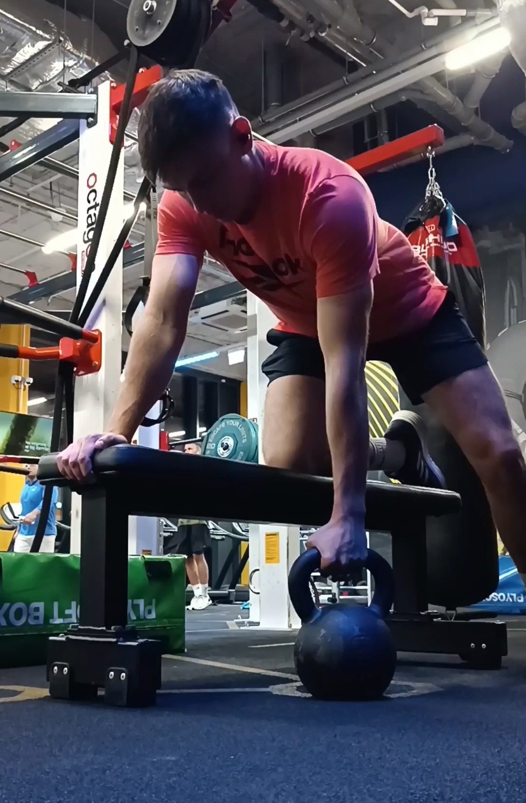 Mężczyzna w czerwonej koszulce wykonuje ćwiczenie z kettlebell na ławce w siłowni, widoczny sprzęt do ćwiczeń i osoby w tle.