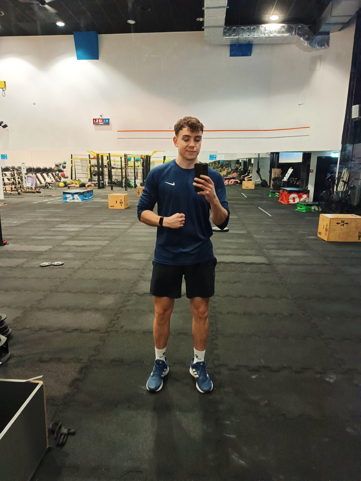 Mężczyzna w sportowym stroju robi selfie w siłowni, w tle widoczne sprzęty do ćwiczeń i osoby trenujące.