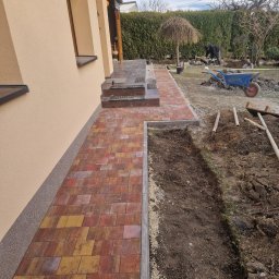 Brukarz - Ścieżka z wielobarwnej kostki prowadząca od domu, z widocznym wykopem pod kolejne prace brukarskie i pracownikiem w tle.